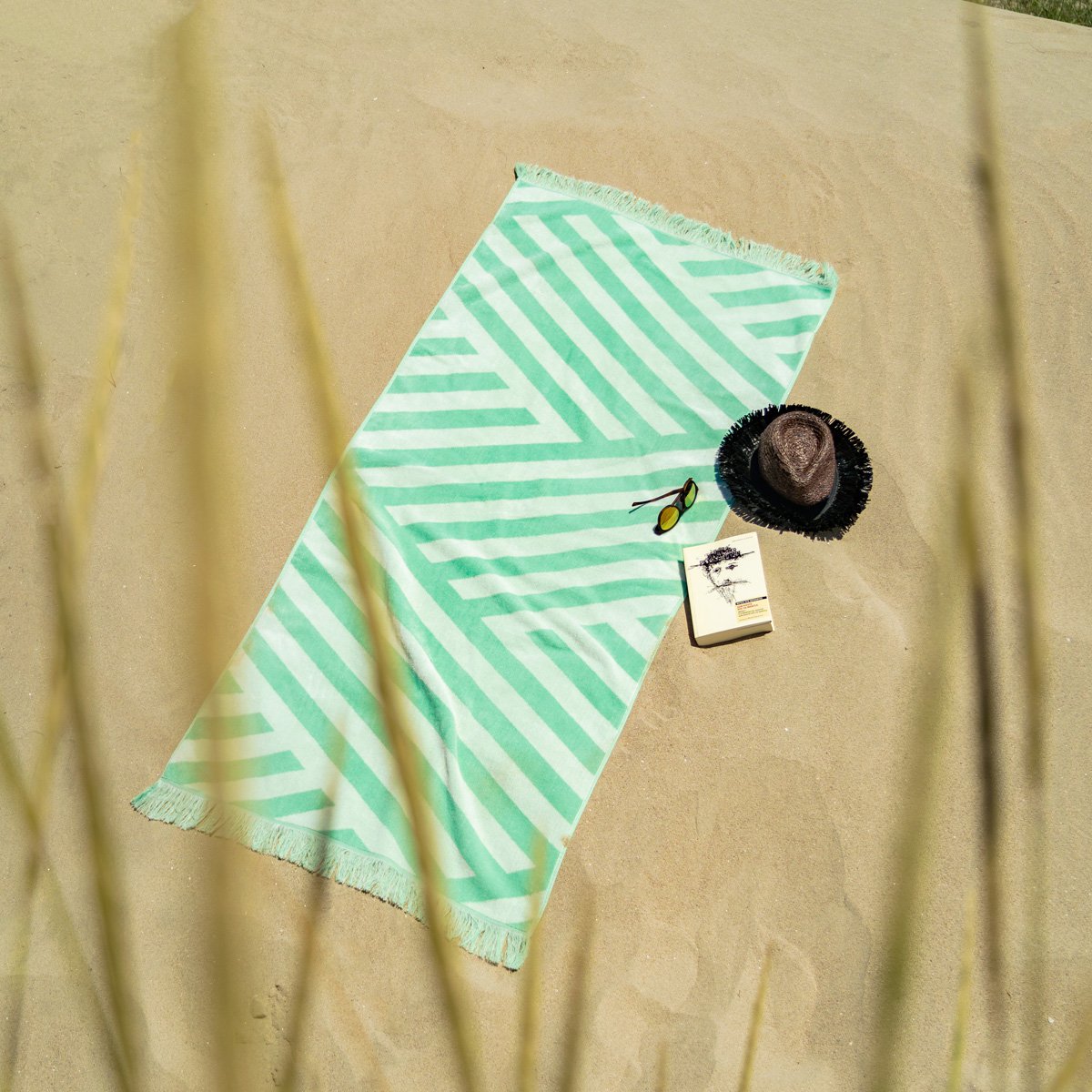 Velours Strandhanddoek Groen met franjes, liggend op het strand naast zonnehoed, boek en zonnebril, perfect voor zonnebaden.