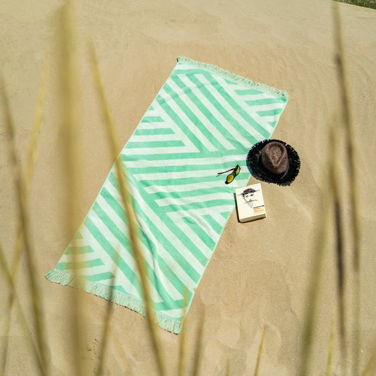 Velours Strandhanddoek Groen met franjes, liggend op het strand naast zonnehoed, boek en zonnebril, perfect voor zonnebaden.