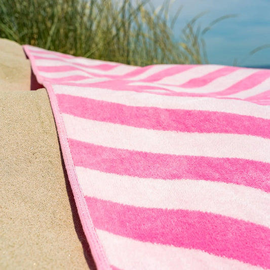 Strandhanddoek Roze met roze-witte strepen van zacht velours, uitgestrekt op zandstrand met gras op de achtergrond.