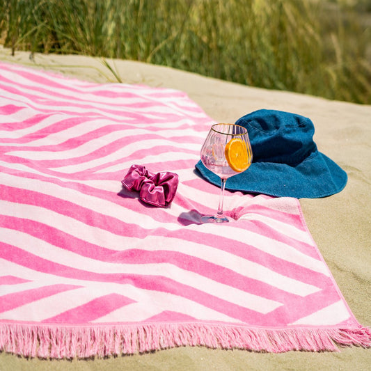 Strandhanddoek Roze met franjes, liggend op strand met accessoires zoals een blauwe hoed, scrunchie en cocktailglas.