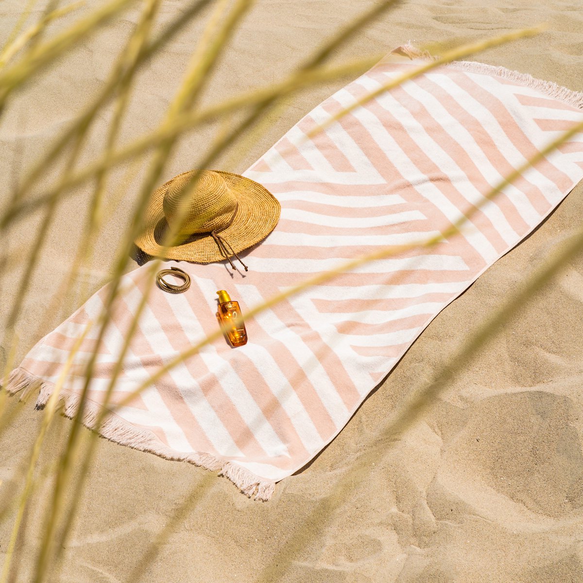 Zachte velours Strandhanddoek Zand in beige-witte strepen, uitgestrekt op het zand met accessoires voor een stranddag.
