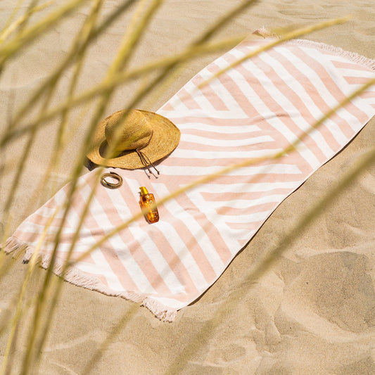 Zachte velours Strandhanddoek Zand in beige-witte strepen, uitgestrekt op het zand met accessoires voor een stranddag.